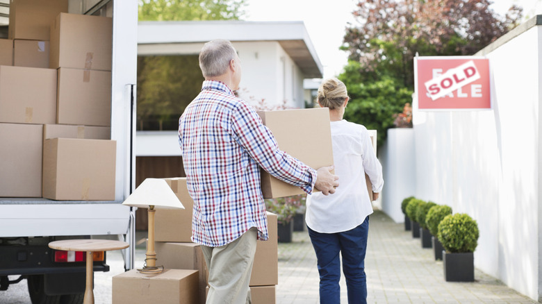 a couple hauls boxes out of a moving van past a for sale/sold real estate sign