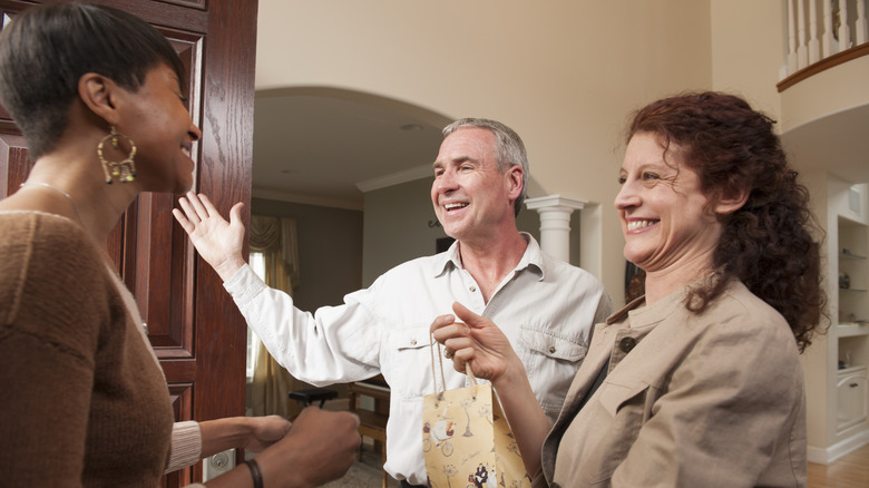A happy couple welcomes a house guest entering through a wood door
