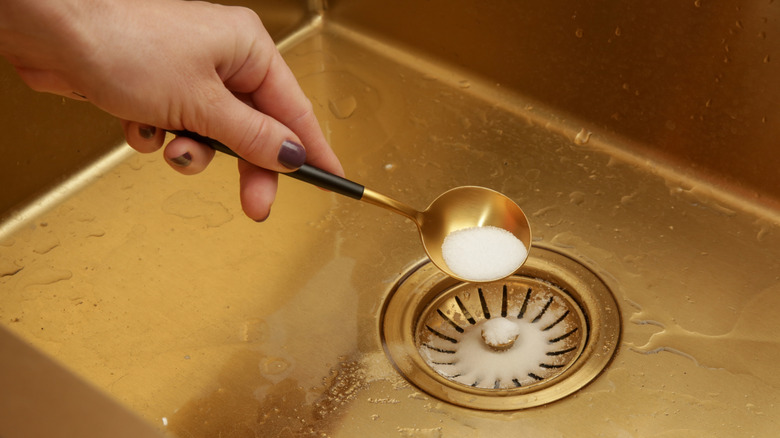 A hand is seen tipping a large spoonful of salt into a sink drain