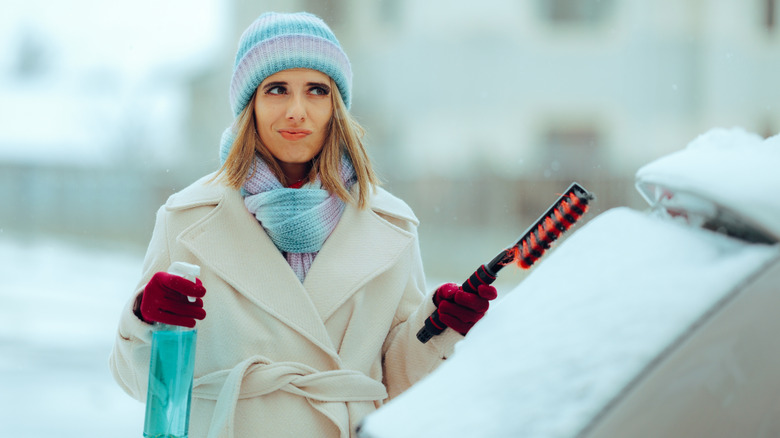 A woman is standing near her snowy car holding a spray bottle and snow brush