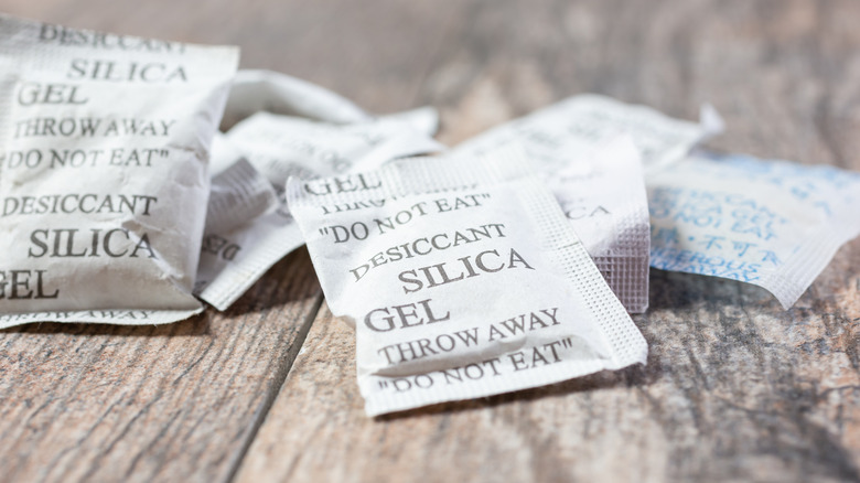 A pile of white silica gel desiccant packets sitting on a greyish wood surface