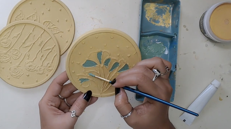 A woman adds teal paint to gold plastic lids decorated with hot glue patterns.