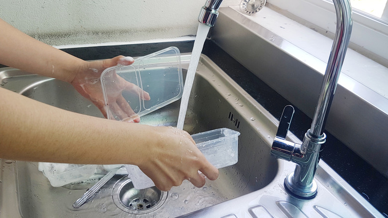 A person cleans an empty plastic takeout container and lid in a stainless steel kitchen sink.
