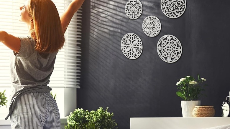 White and black medallion art hangs on a wall in a bedroom while a lady faces the window and stretches.