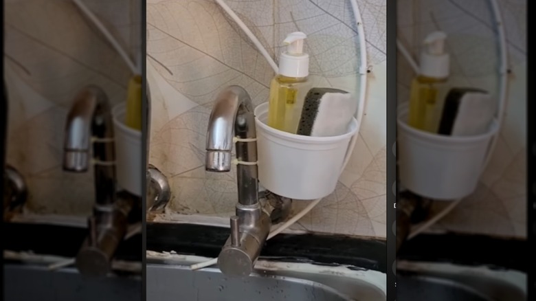 A sponge holder tied to the side of the faucet made from a plastic container and zip ties.