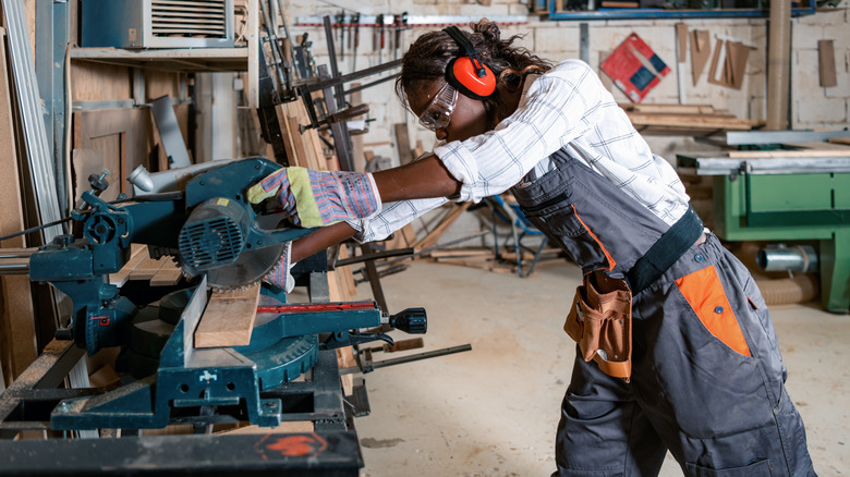 Woman wearing earmuffs and goggles cutting piece of wood with table saw in workshop
