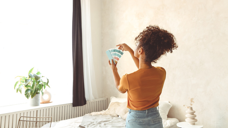 A woman chooses a paint color while standing in front of her bed