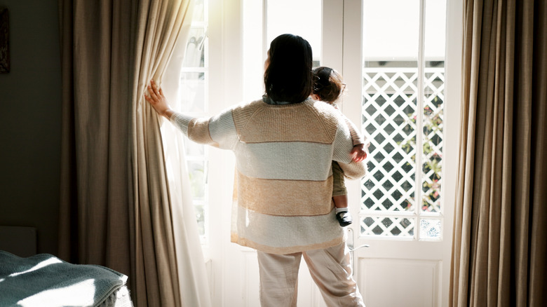 A woman standing in front of a window with her baby, pulling back the curtains.