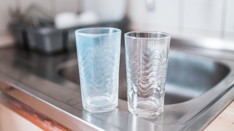 One clean and one cloudy glass tumbler sit on the edge of a stainless steel kitchen sink.