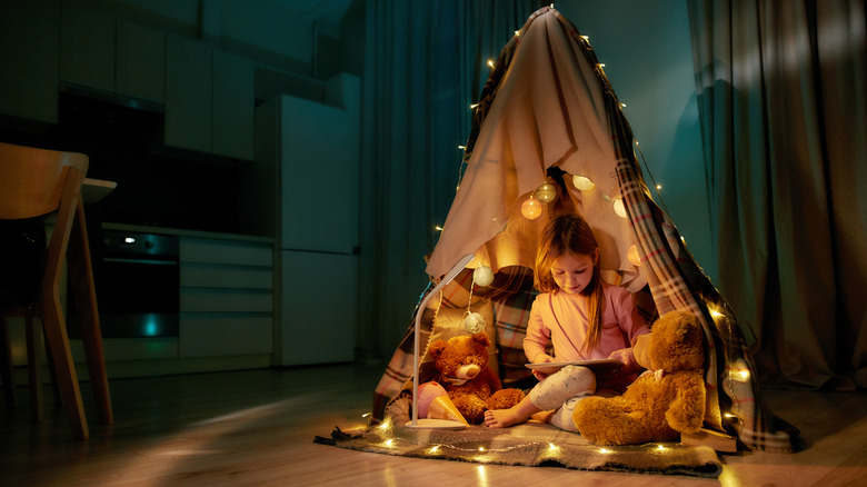A child reading a tablet in a tent with string lights and teddy bears