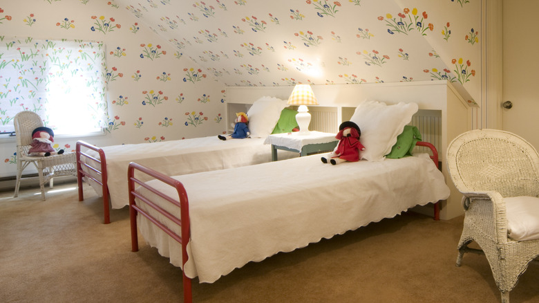 Two twin beds with dolls on them in a room with floral wallpaper and wicker chairs