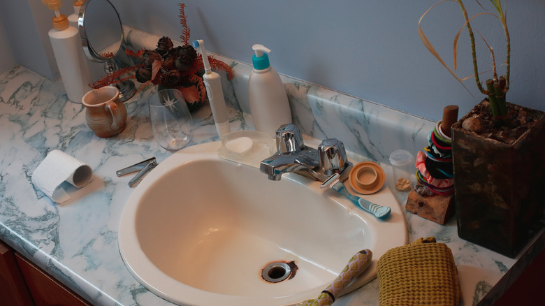 A bathroom looks messy when there are a lot of toiletries on the sink top in addition to an almost-empty roll of toilet paper, a glass, a mug, a mostly-dead plant, and other items.