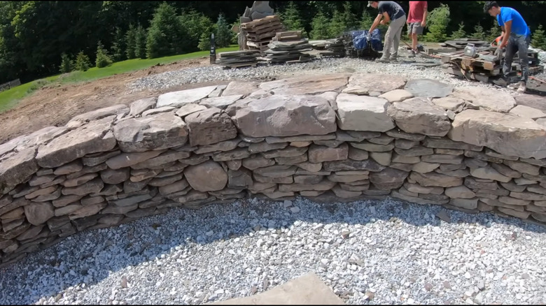 A dry stack stone retaining wall with gravel on one side and people working nearby
