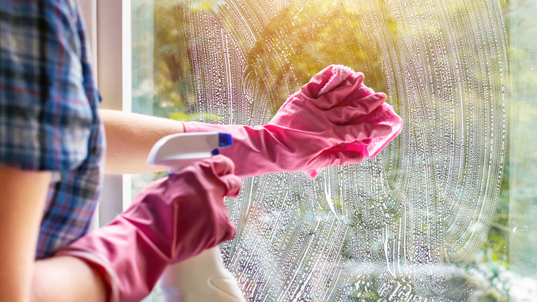 A woman wearing pink cleaning gloves scrubs a window with a sponge