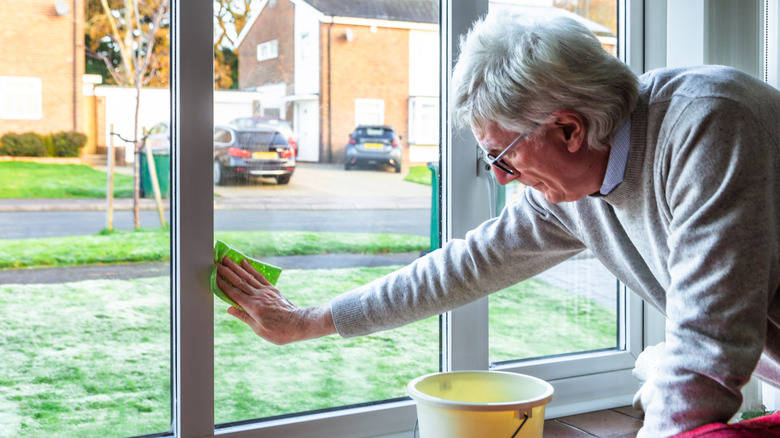 A senior man wipes down front windows with a green sponge.