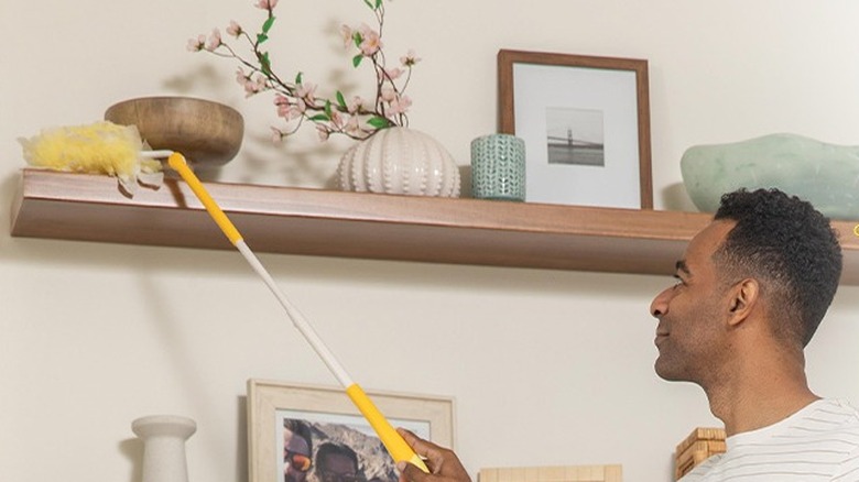 A man using a yellow and white Swiffer duster to clean a shelf