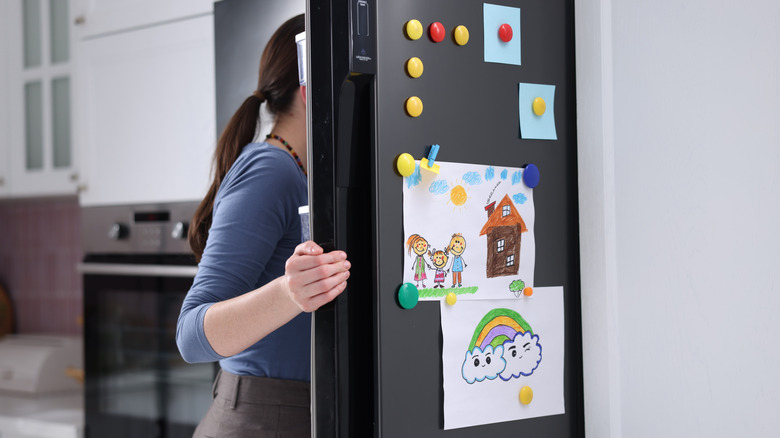 Person opening refrigerator with child's drawings on it indoors, closeup