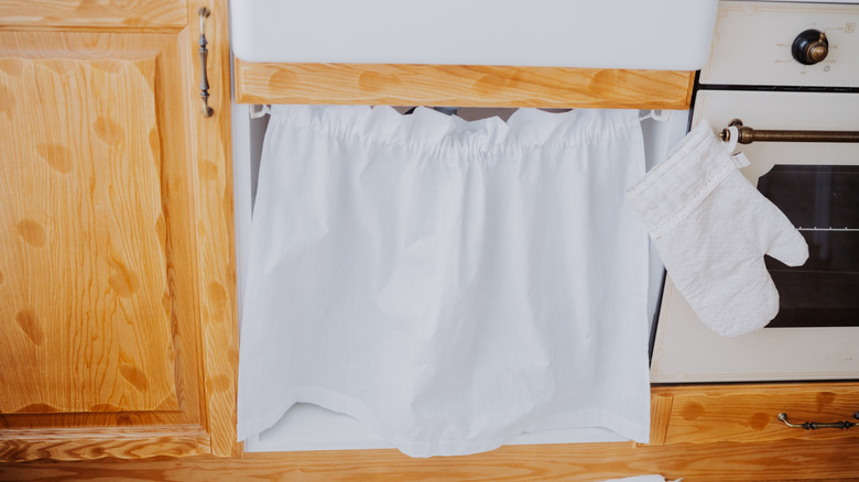 A white curtain under a sink in a kitchen next to a stove and a cabinet