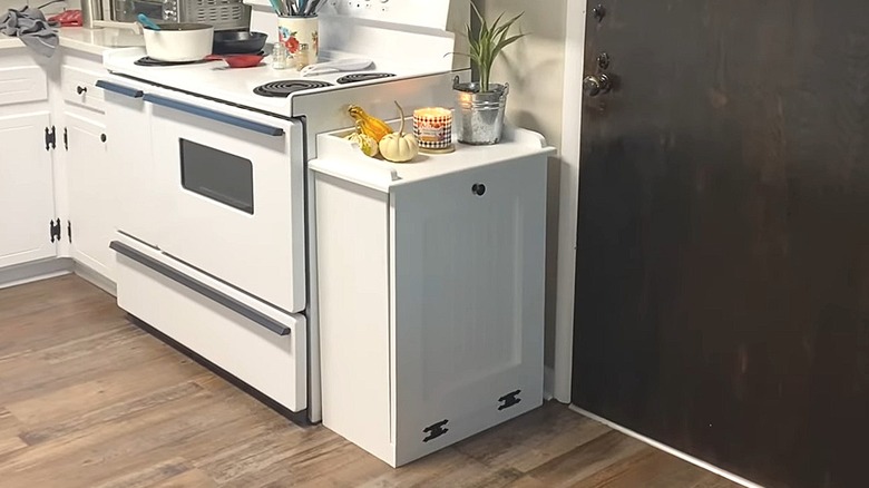 White wooden tilt-out trash can cabinet next to a stove with decor sitting on top