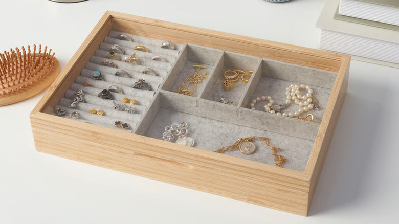 The HALSREM jewelry box with compartments full of rings and necklaces of different materials on a counter next to a brush and some books