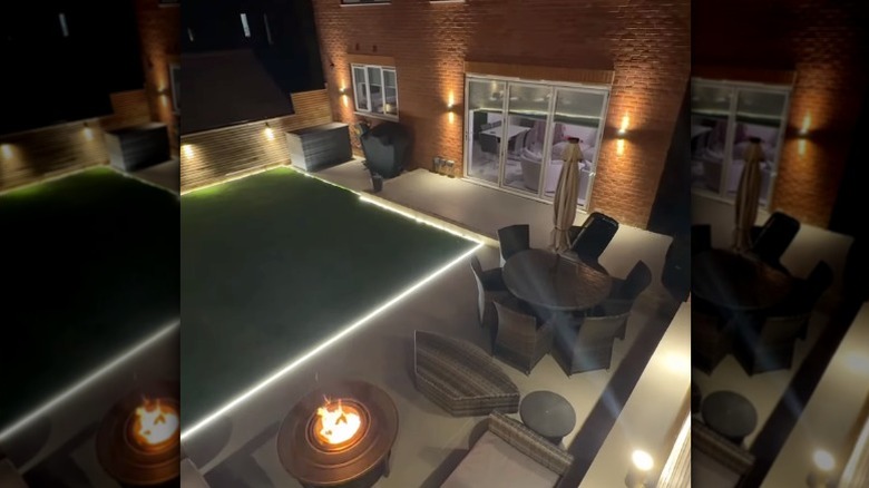 A backyard patio with outdoor furniture and a firepit illuminated by lights.
