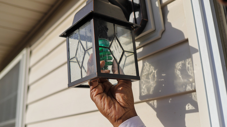 A person changes the lightbulb in a traditional wall-mounted outdoor light fixture