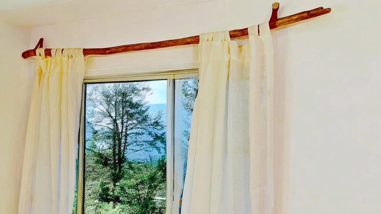 Curtains hang from a driftwood branch over a window with a view outside to large trees.