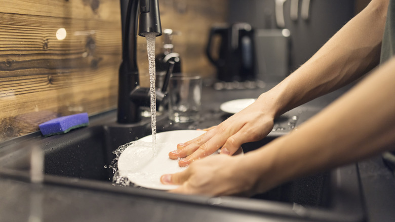 A person runs water over a white plate while washing it in the kitchen sink.