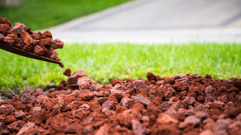 A pile of red lava rock in front of a lawn with more lava rock in the bowl of a shovel hovering above the pile.