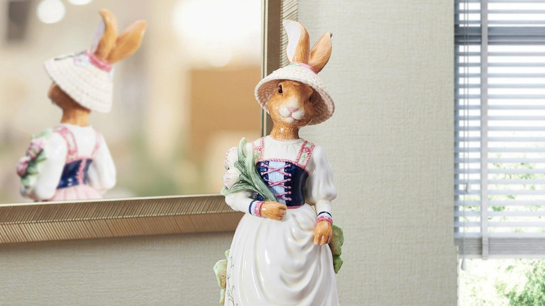 A well-sculpted figurine of a rabbit in a delicate dress is perched on a book atop a table near a mirror