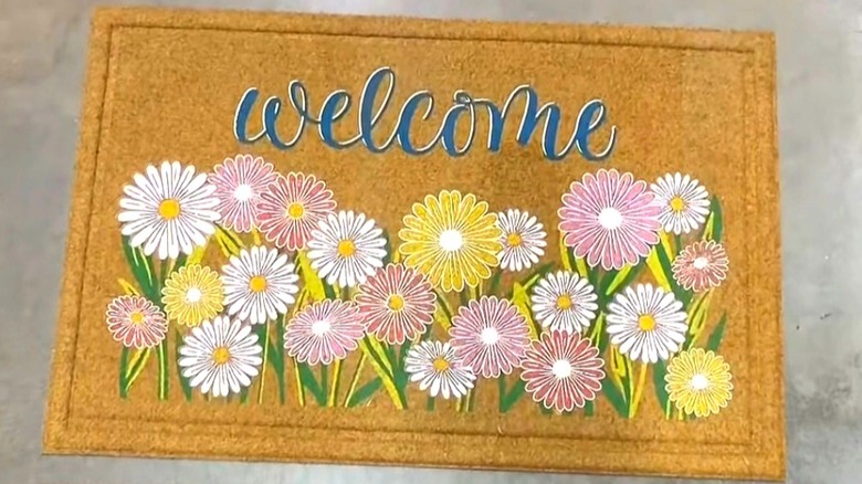 A decorative door mat with the word "Welcome" in script above a floral design sits on a warehouse floor
