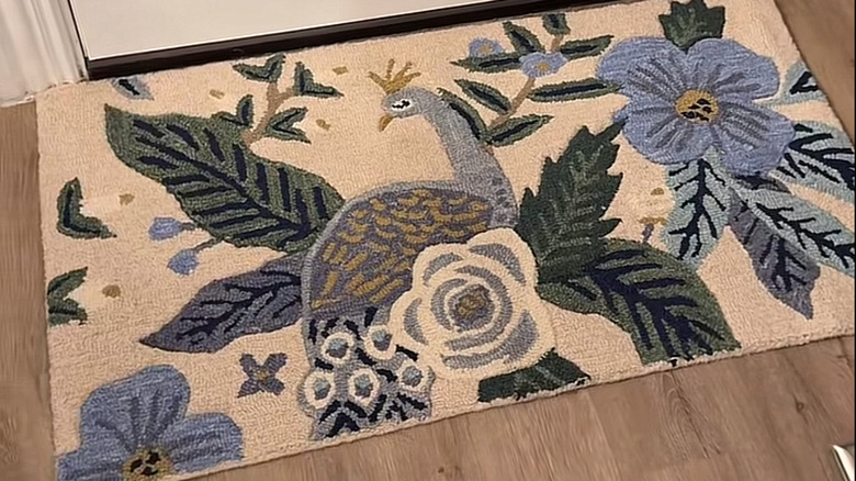 A rug with a peacock and botanical print sits on a wooden floor in front of a white door