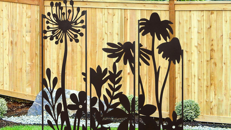 A dark steel outdoor mural stands in front of a wooden fence and trees behind it