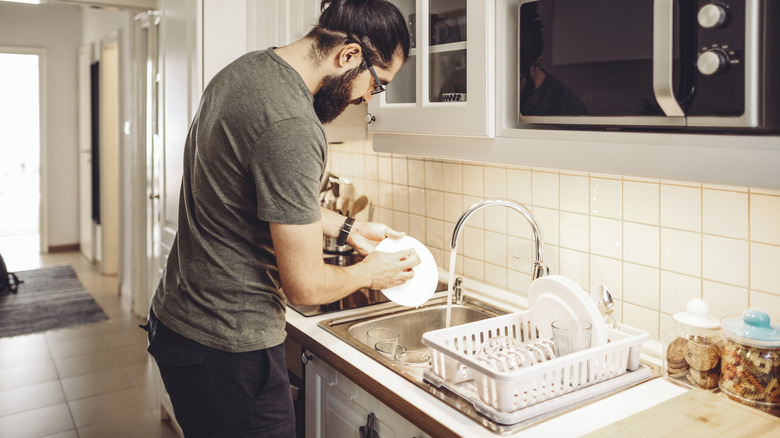 A man washes dishes in a kitchen sink using a sponge