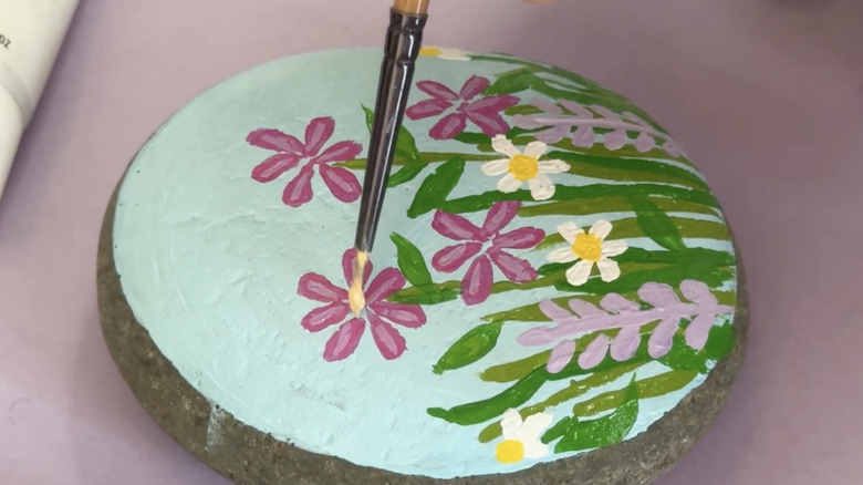 A person uses a paintbrush to paint flowers on a smooth round river rock.
