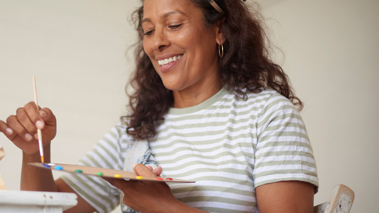 A smiling woman sits at a table holding a paintbrush and paint palette.