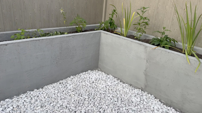 A concrete flower bed filled with plants sits in a fenced corner of a backyard lined with white gravel.