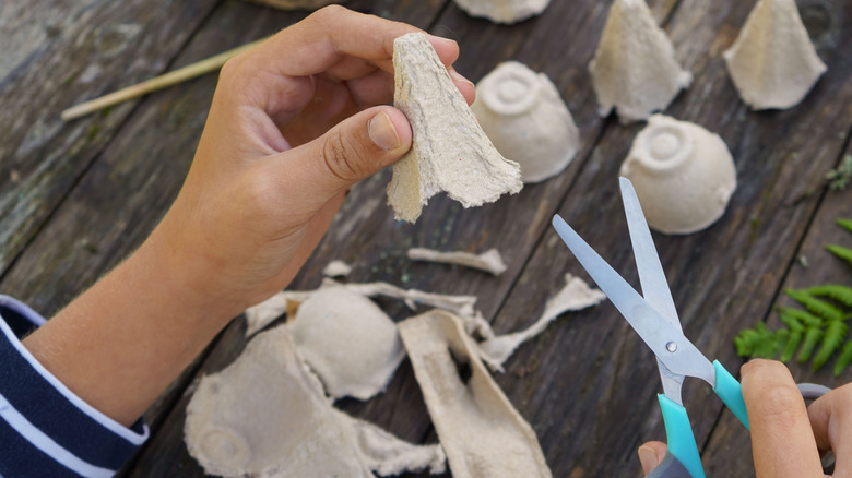 A person holds an egg carton cup in one hand and a pair of scissors in the other while working at a wooden table outside.
