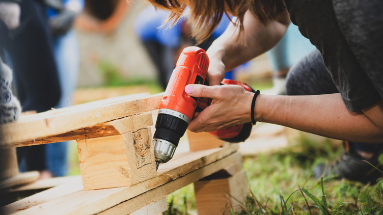Woman using a power drill on a wooden pallet, which is laying in the grass.
