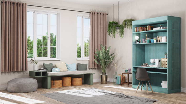 A large open living room with a shelving unit and work station painted a bright aqua color to contrast with the white walls, dusky pink curtains and collection of plants