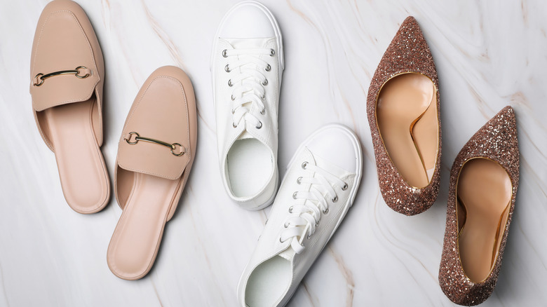 Three pairs of shoes on a marble floor: beige mules, white sneakers, and sparkly high heels.