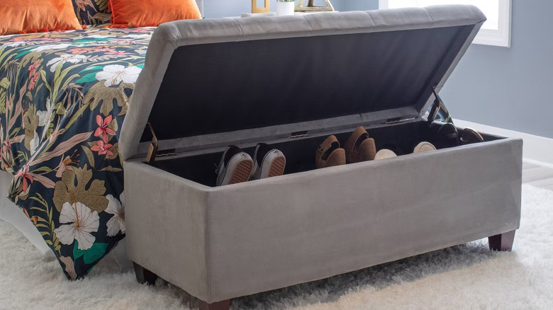 A grey ottoman filled with shoes sitting at the end of a bed with dark green floral bedding and orange cushions.