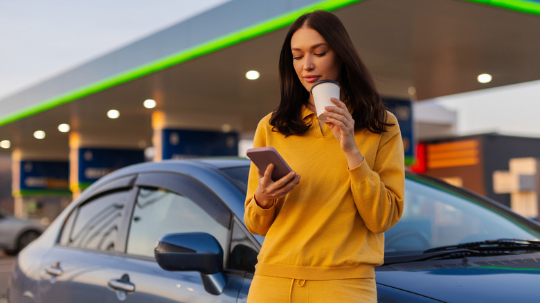 Woman checking her phone drinking a coffee leaning on a car door at a gas station