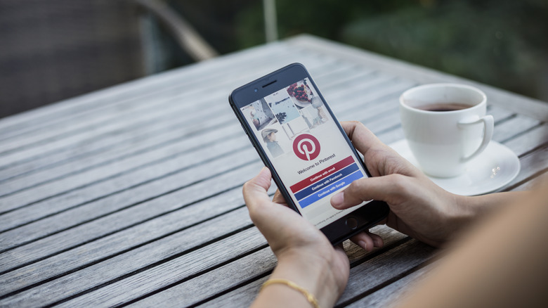 Person using Pinterest on their phone while sitting at a table with a cup of coffee on a saucer