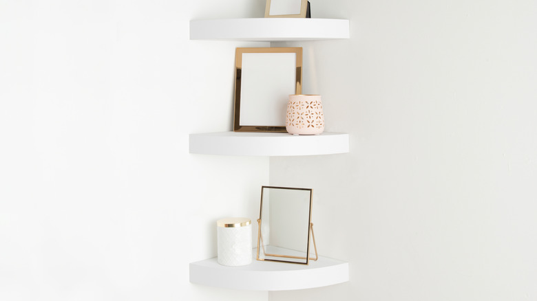 Corner nook with sleek white floating shelves, gold-framed mirrors, pink cutout candle holder, and a glossy white jar.