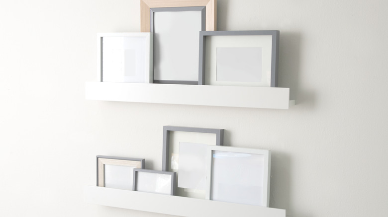 Two floating white ledges mounted on light wall, displaying assorted empty photo frames in white, gray and natural wood tones.