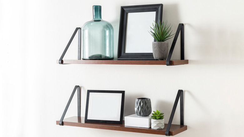 Scandinavian inspired composition featuring two floating wooden shelves with black metal brackets, decorated with framed pictures, green glass bottle, mini succulents and ceramic vases on white wall.