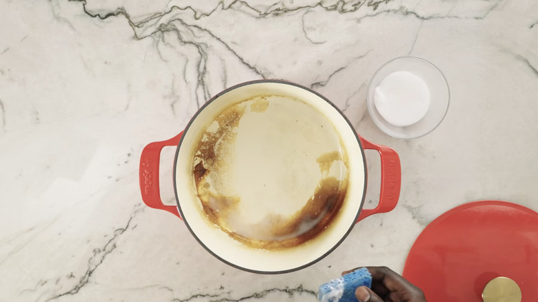 A person cleans a red enameled cast iron pot with vinegar, baking sofa, and a blue sponge.