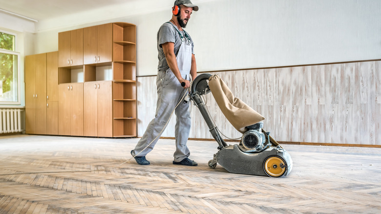 A man in a hat and overalls pushing a sanding machine along a hardwood floor.