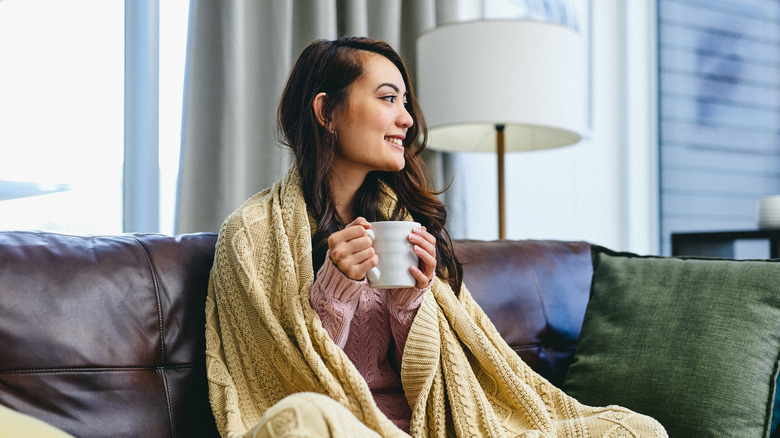 Woman wrapped in throw blanket and holding hot drink on couch